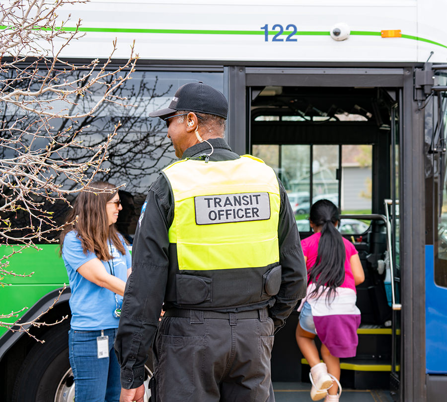 A transit officer standing on the curb smiles as riders get on a bus.