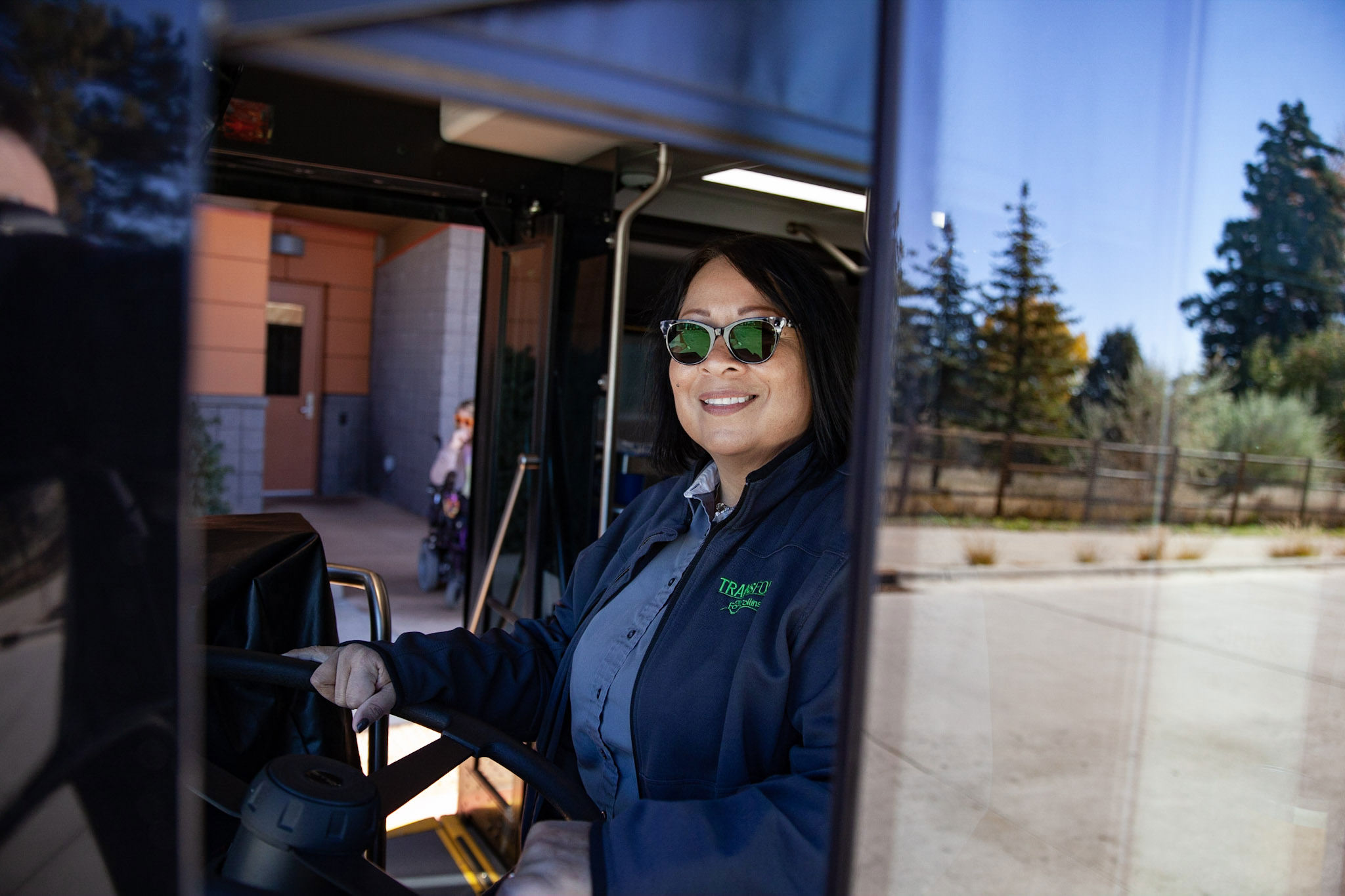 A bus driver in sunglasses smiles in the driver's set of a bus. 